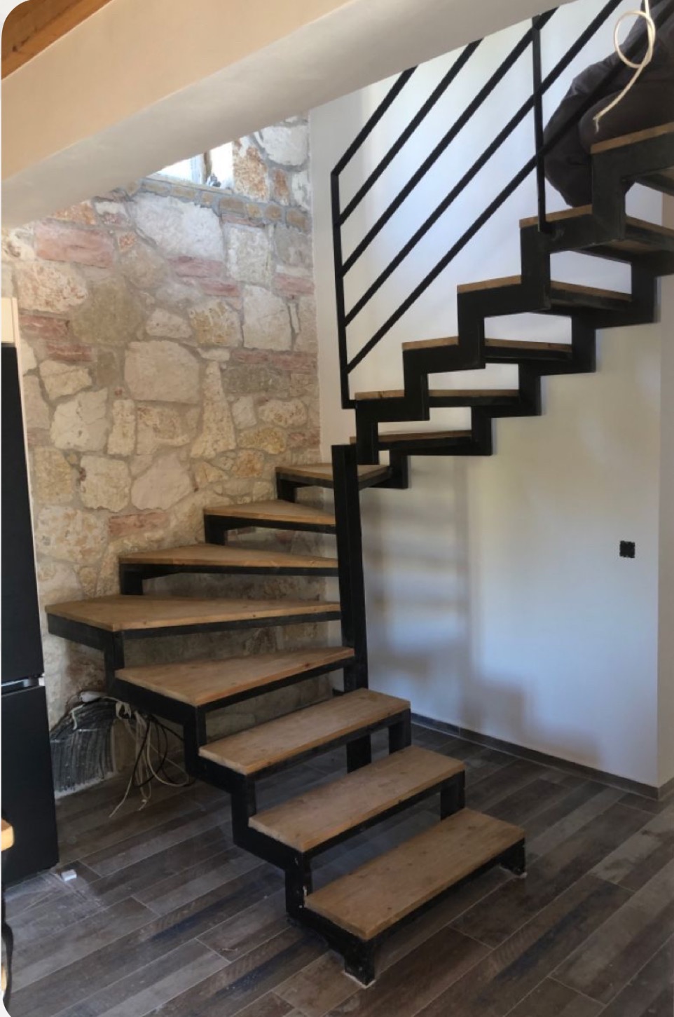Nowoczesne schody metalowe z drewnianymi stopniami, czarna balustrada, ściana z kamienia w tle. Nietypowy układ stopni, minimalistyczny design.