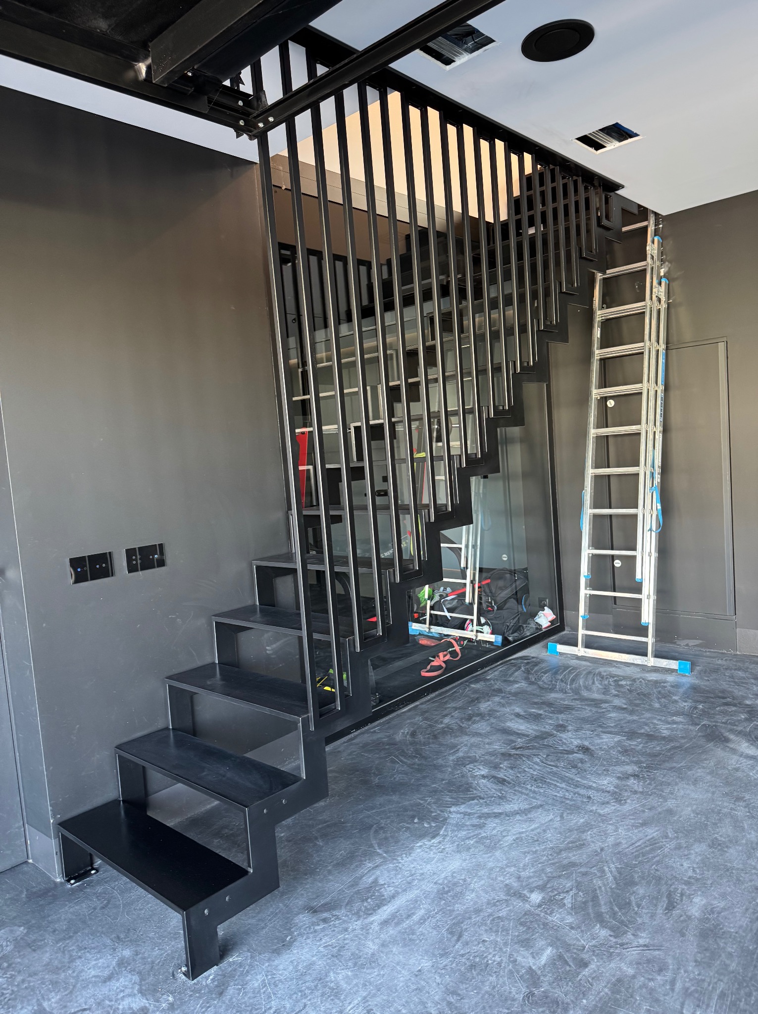 Nowoczesne, czarne schody metalowe z minimalistyczną balustradą w industrialnym wnętrzu. Widoczna drabina i narzędzia sugerują etap montażu.