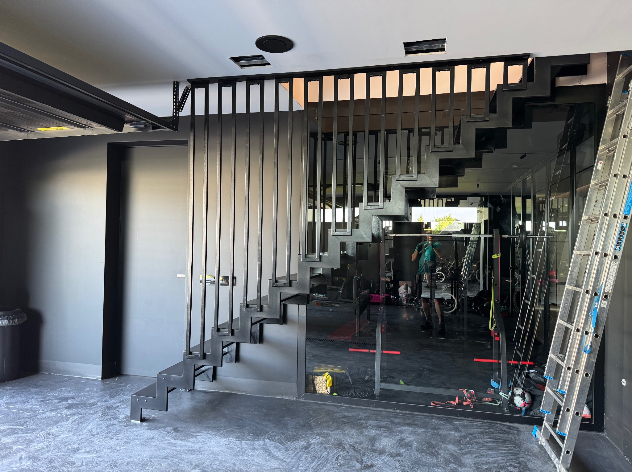 Nowoczesne, czarne schody metalowe z minimalistyczną balustradą, widoczne w przestronnym, ciemnym wnętrzu. Aluminiowa drabina oparta o ścianę. Lustrzane wykończenia w tle.