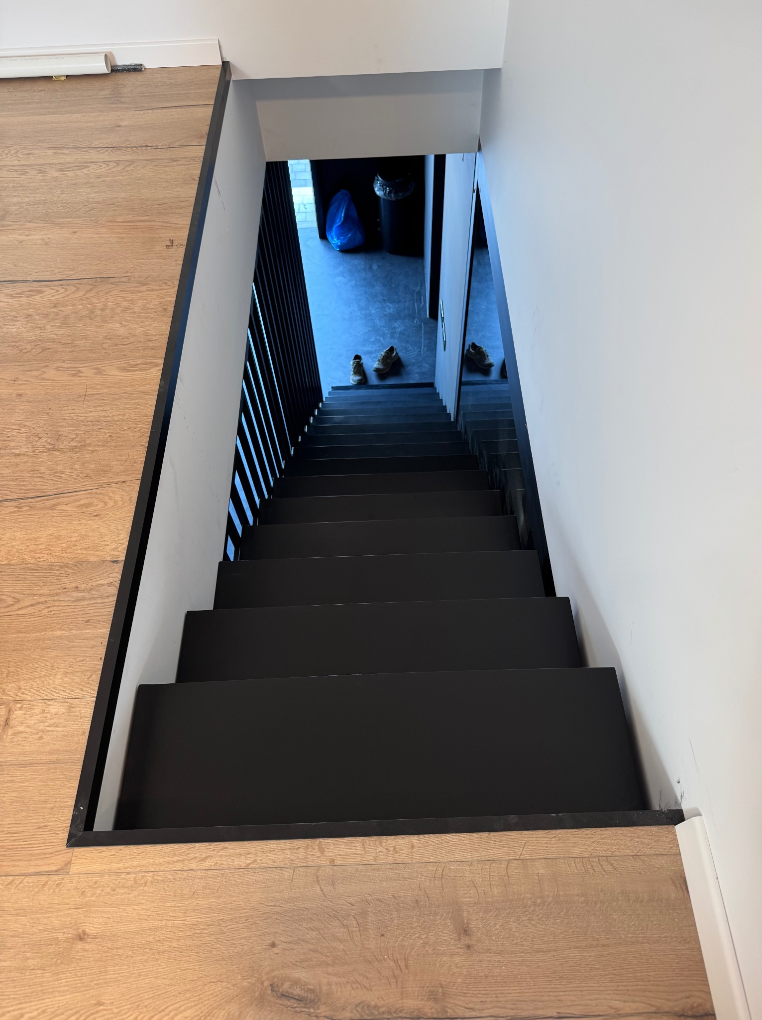 Nowoczesne schody w dół z czarnymi stopniami i minimalistyczną balustradą, widziane z góry. Jasne drewno podłogi kontrastuje z bielą ścian.