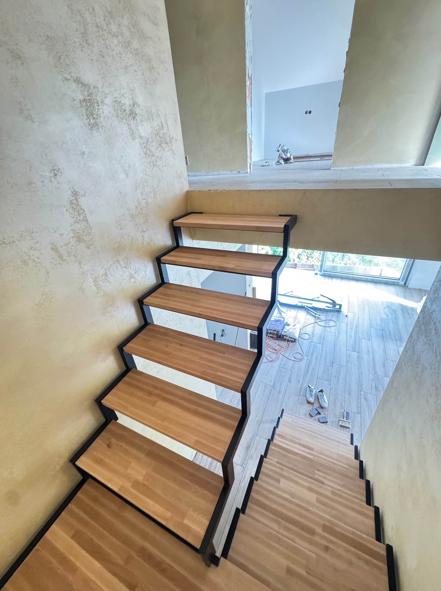 Nowoczesne schody z drewnianymi stopniami i czarną metalową konstrukcją, widok z góry, w tle okno i narzędzia, surowe ściany w beżowym odcieniu.