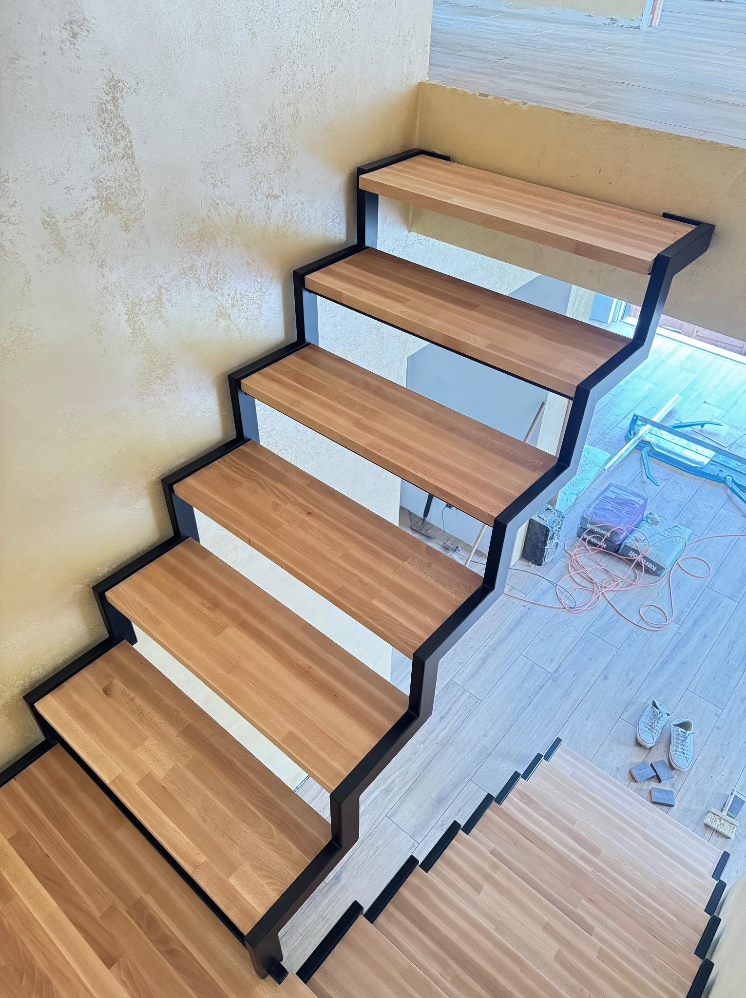 Nowoczesne schody z drewnianymi stopniami i czarną, metalową konstrukcją, widziane z góry. Na podłodze leżą narzędzia i buty robocze.