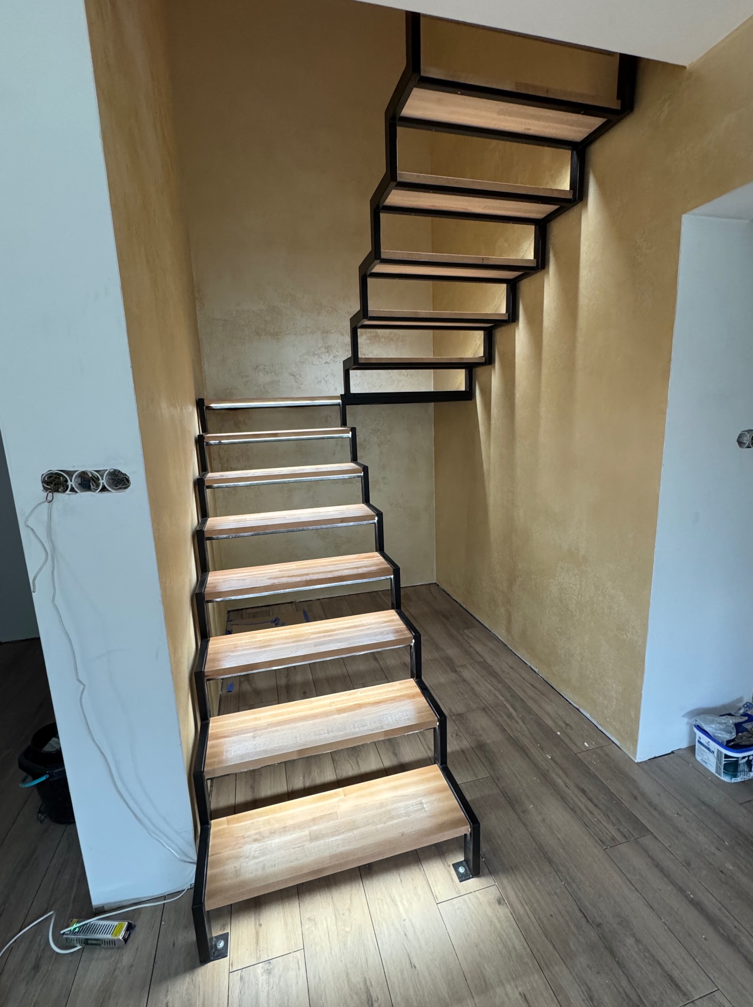 Nowoczesne schody z drewnianymi stopniami i czarną metalową konstrukcją w jasnym wnętrzu. Widoczne gniazdka elektryczne i faktura ściany.