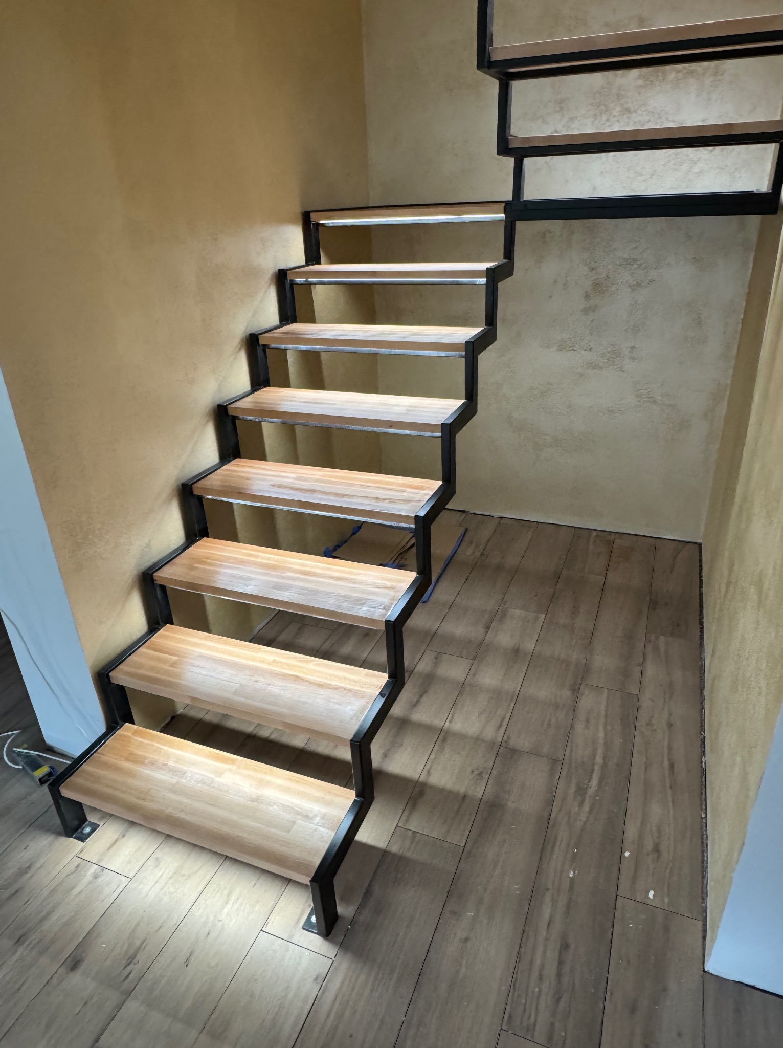 Nowoczesne schody drewniane z czarną, metalową konstrukcją w jasnym wnętrzu. Widoczne drewniane stopnie i dekoracyjna ściana w odcieniach beżu.