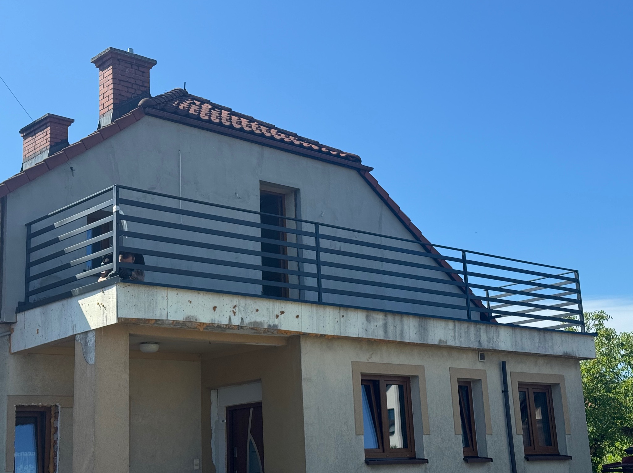 Dom z balkonem zabezpieczonym nowoczesną, poziomą, czarną barierką. Ceglany komin na dachu i okna z brązowymi ramami dopełniają architekturę.