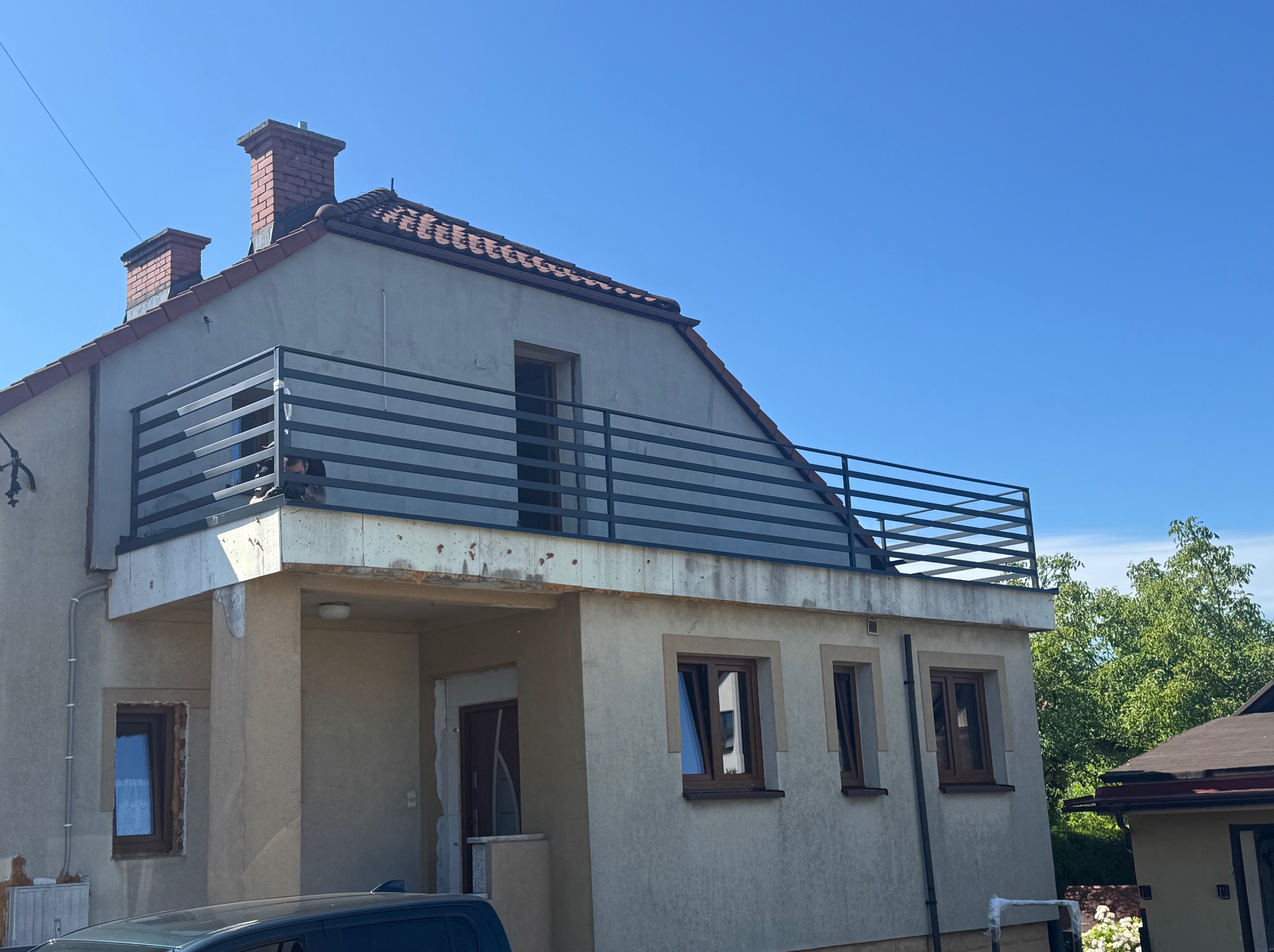 Dwukondygnacyjny dom w trakcie remontu z czarną, poziomą balustradą na balkonie. Widoczne okna z brązowymi ramami i fragment samochodu na pierwszym planie.