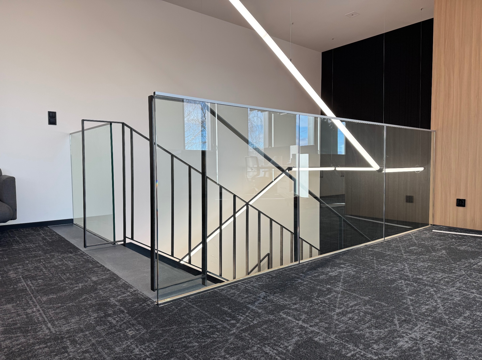 Nowoczesne schody z balustradą szklaną i czarnymi poręczami, widok z góry na szary dywan. Minimalistyczny design, oświetlenie LED. Widoczne odbicie drzew w szkle.
