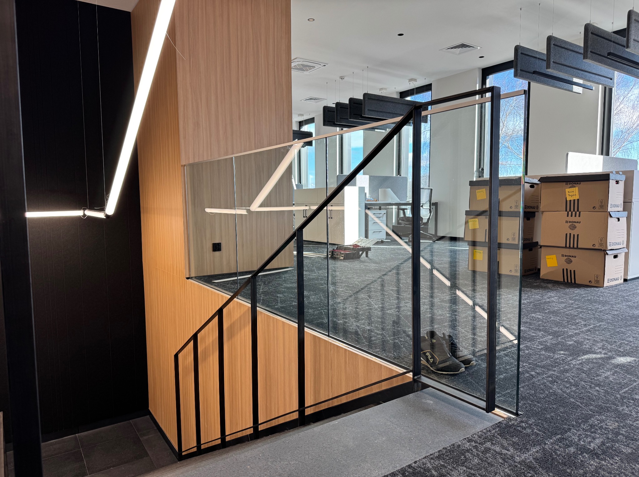 Nowoczesne schody z balustradą szklaną i czarnymi elementami metalowymi w jasnym biurze. Widoczne kartony i oświetlenie sufitowe.