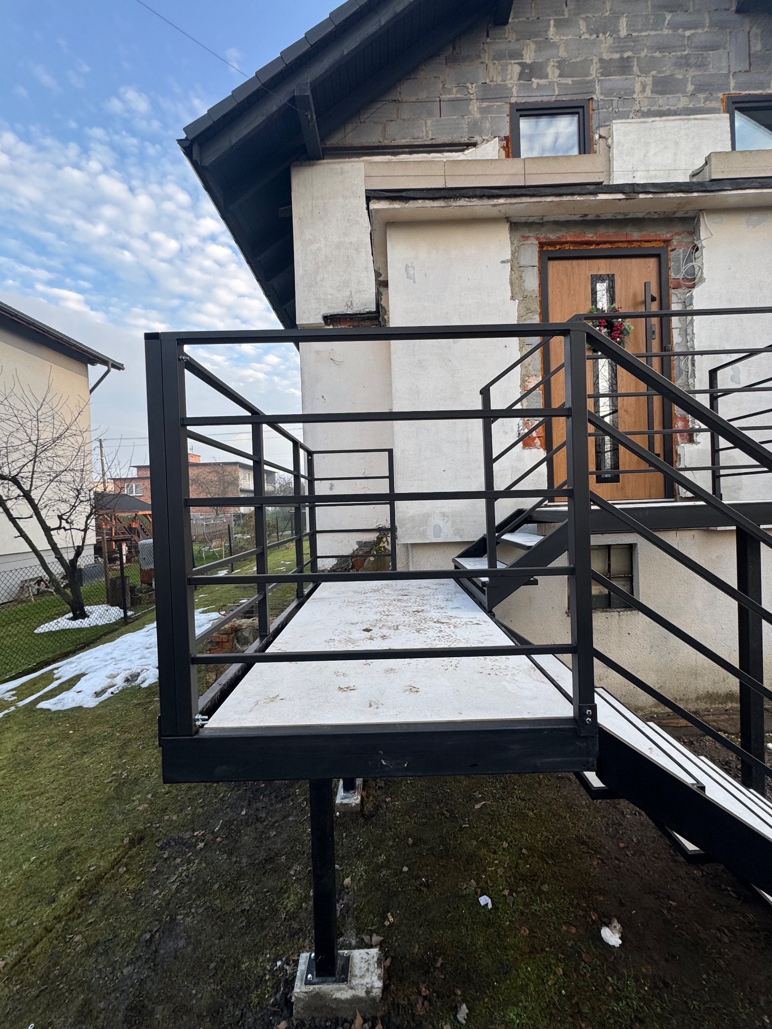Czarna, metalowa konstrukcja schodów zewnętrznych z balustradą, prowadząca do drzwi wejściowych domu w trakcie budowy. Widoczne elementy mocujące i surowy stan elewacji.