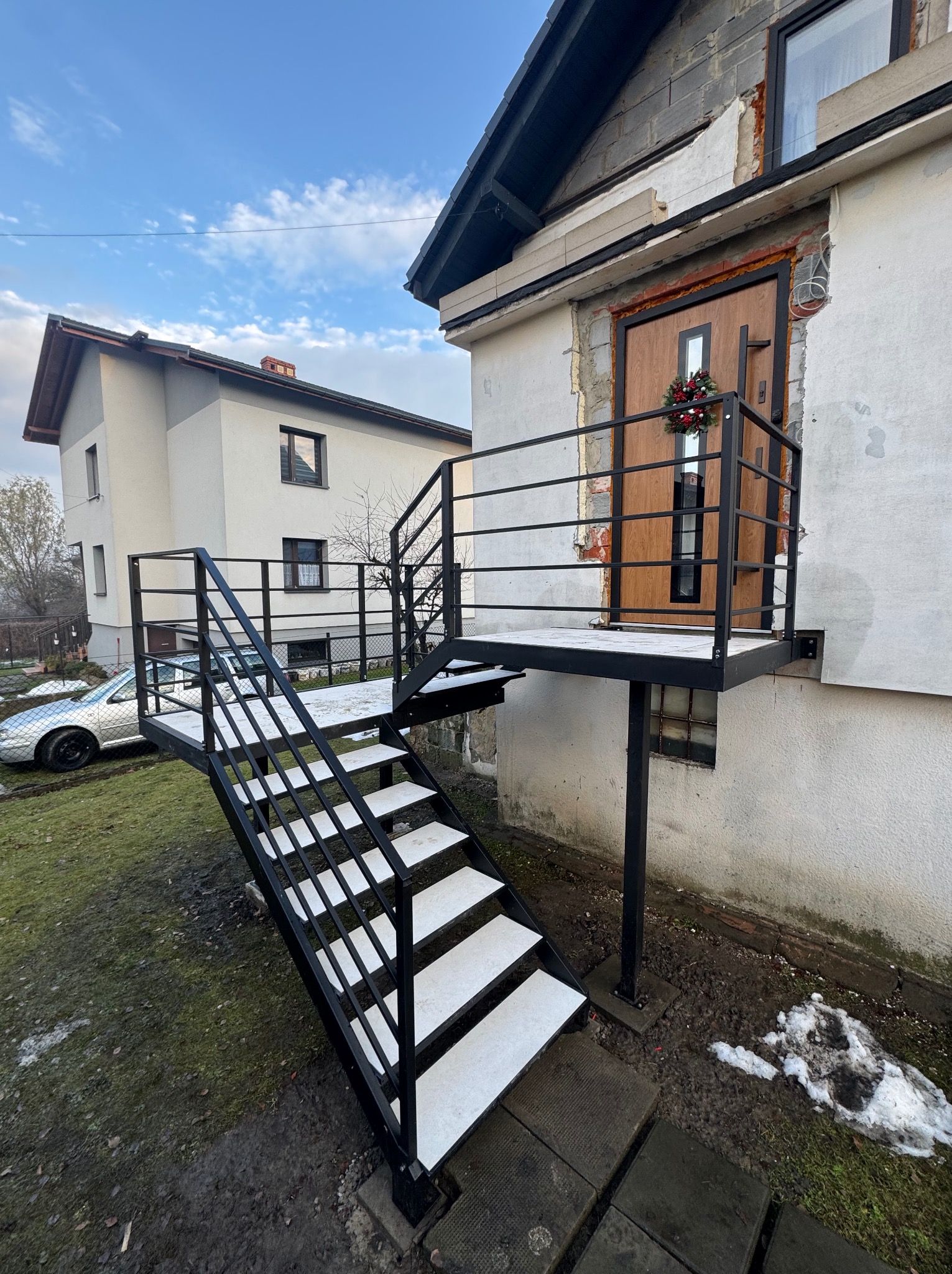 Zewnętrzne schody metalowe z białymi stopniami prowadzące do drzwi wejściowych z balustradą. Widoczne niedokończone wykończenie elewacji budynku.