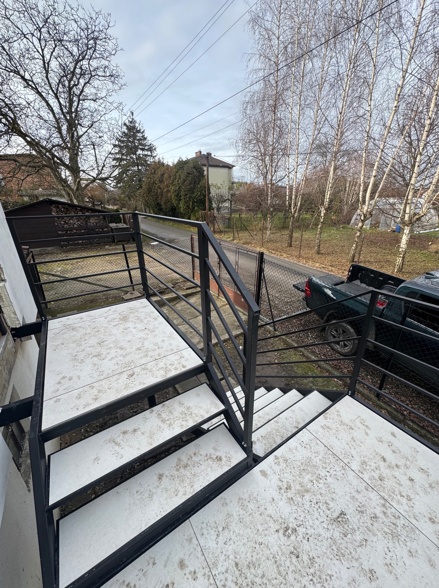Zewnętrzne schody z czarną, metalową balustradą i białymi stopniami, widziane z góry, z elementami otoczenia: drzewa, ogrodzenie i zaparkowany samochód.