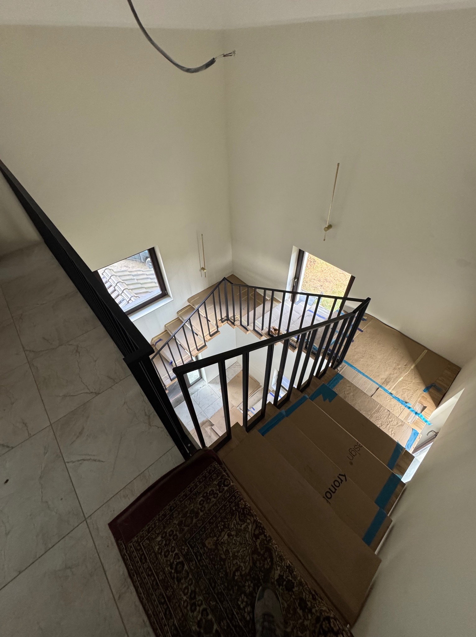 Wysoki kąt: czarna balustrada schodowa z kartonowym zabezpieczeniem stopni. Widoczne okna i fragment dywanu. Minimalistyczny design, jasne ściany.