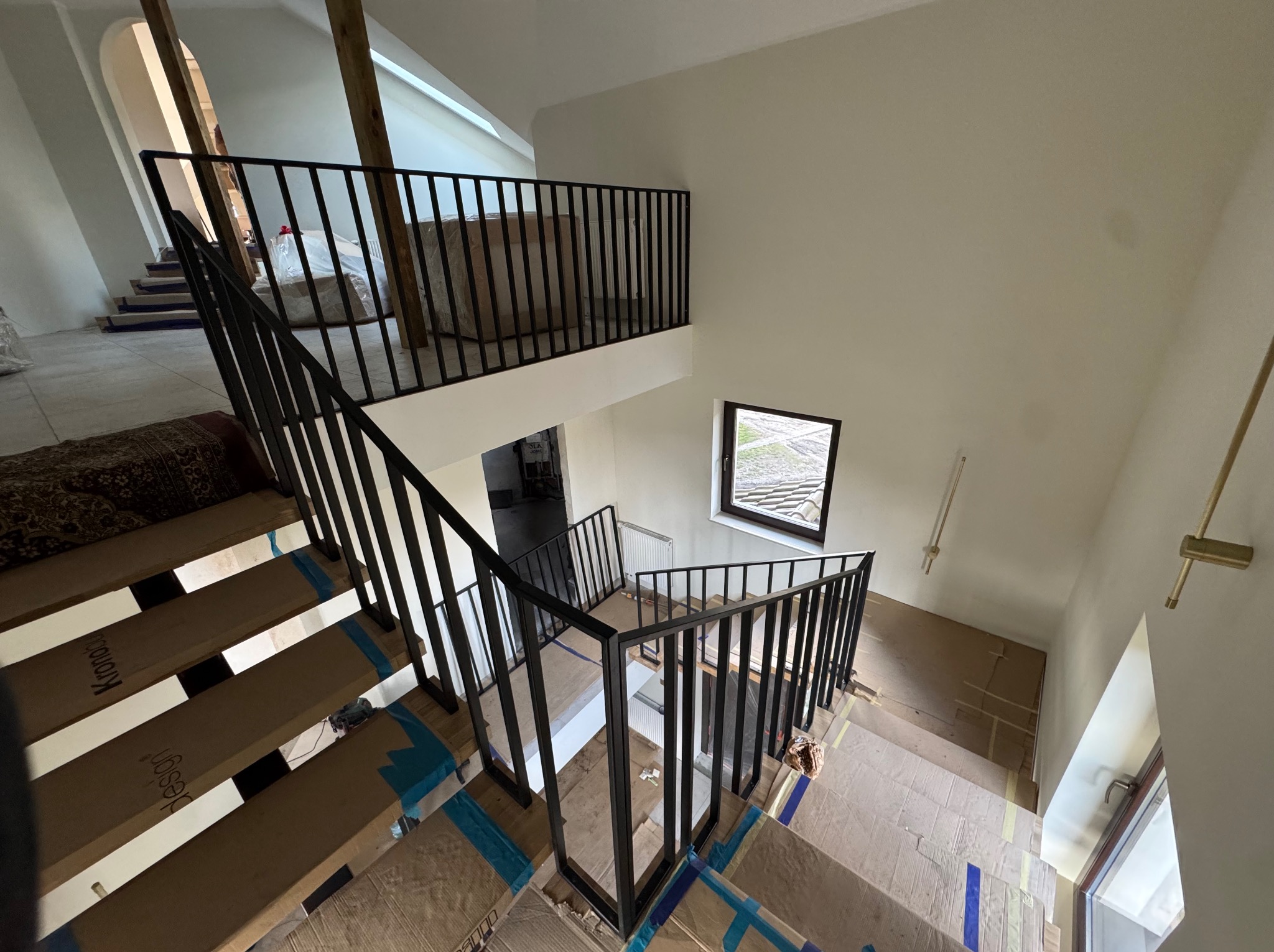 Nowoczesne schody z czarną, metalową balustradą w jasnym wnętrzu. Stopnie zabezpieczone kartonem. Widok z góry na dwupoziomową konstrukcję.