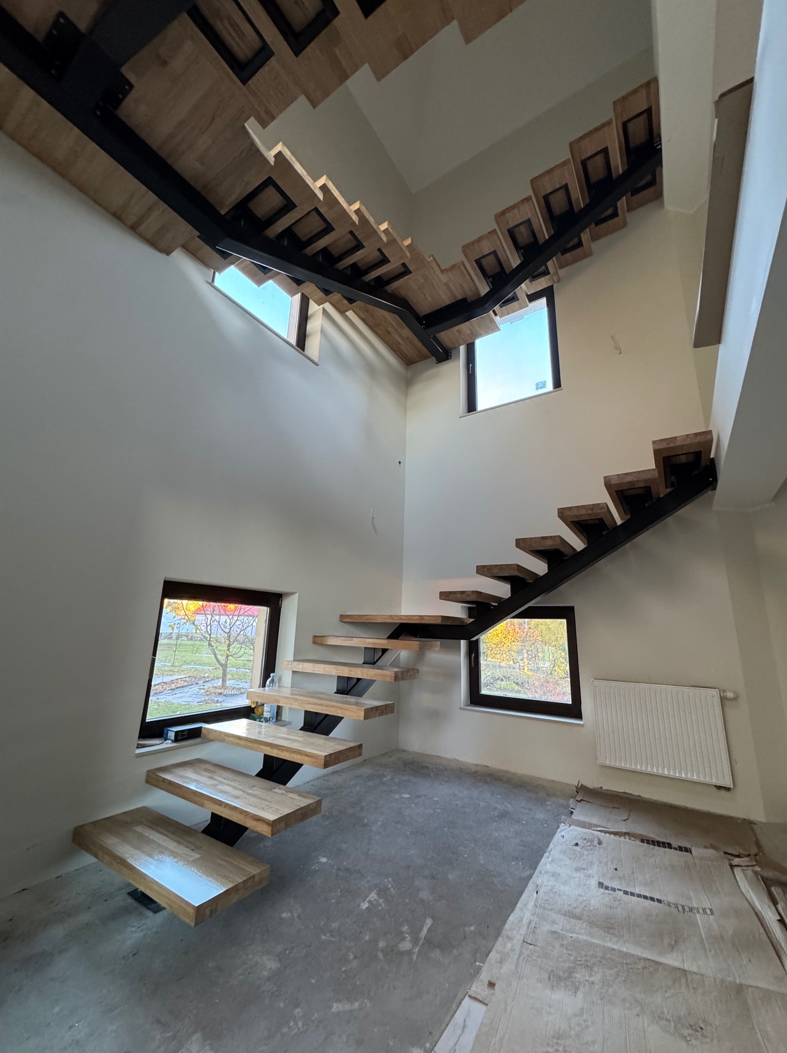 Nowoczesne schody drewniane z metalową konstrukcją w jasnym wnętrzu z oknami. Stopnie z jasnego drewna, czarna metalowa konstrukcja. Widok z dołu na wysokie pomieszczenie.