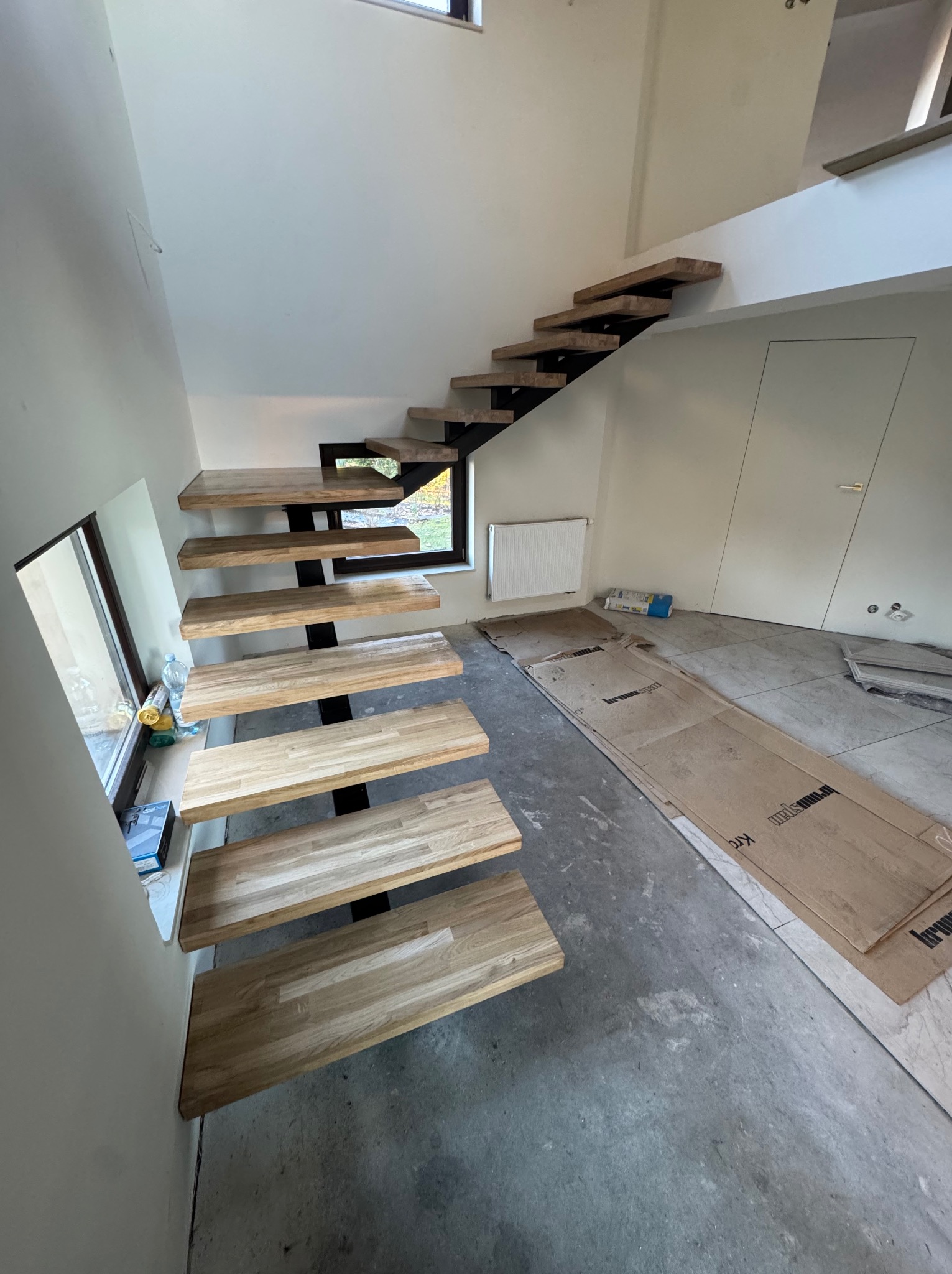 Nowoczesne schody drewniane z metalową konstrukcją w jasnym wnętrzu. Stopnie z jasnego drewna, czarne metalowe wsporniki. Widok z dołu, okno obok schodów.