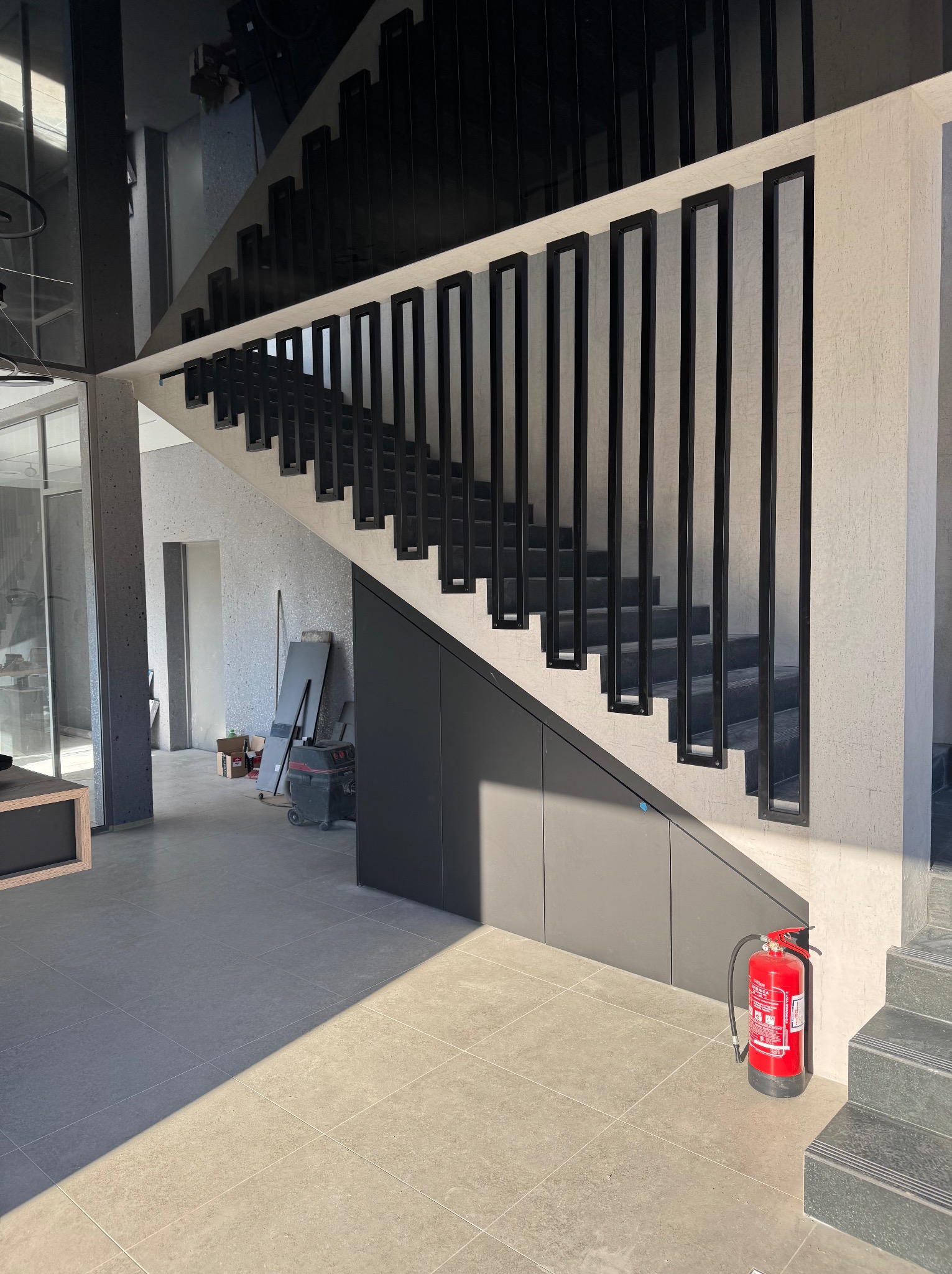 Nowoczesne schody z czarnymi, prostokątnymi balustradami w jasnym wnętrzu. Widoczny fragment pod schodami z zabudową i gaśnica. Minimalistyczny design.