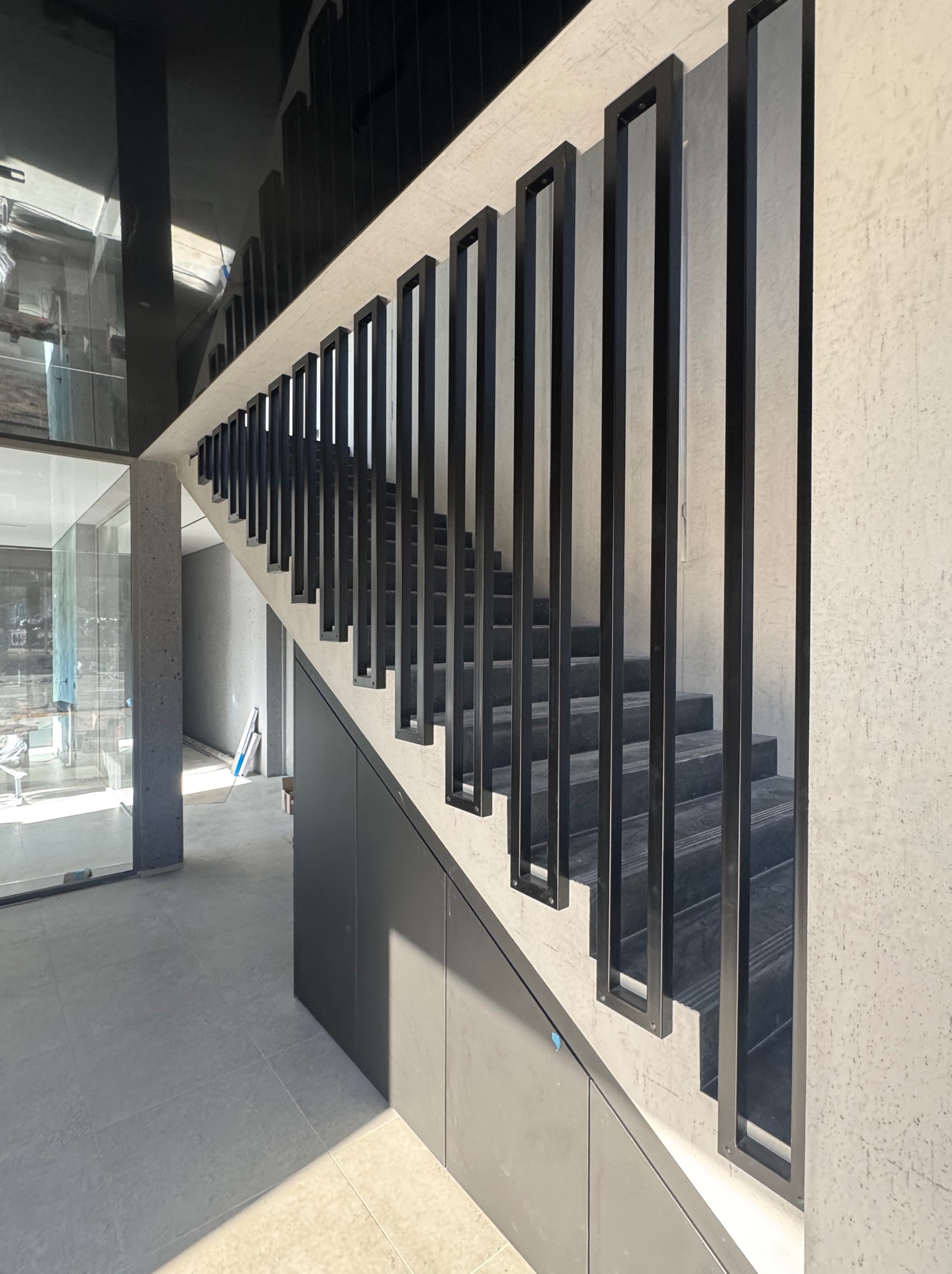 Nowoczesne schody z czarną, minimalistyczną balustradą z prostokątnych elementów. Betonowe wykończenie ścian i podłogi. Widok z dołu, podkreślający geometrię.