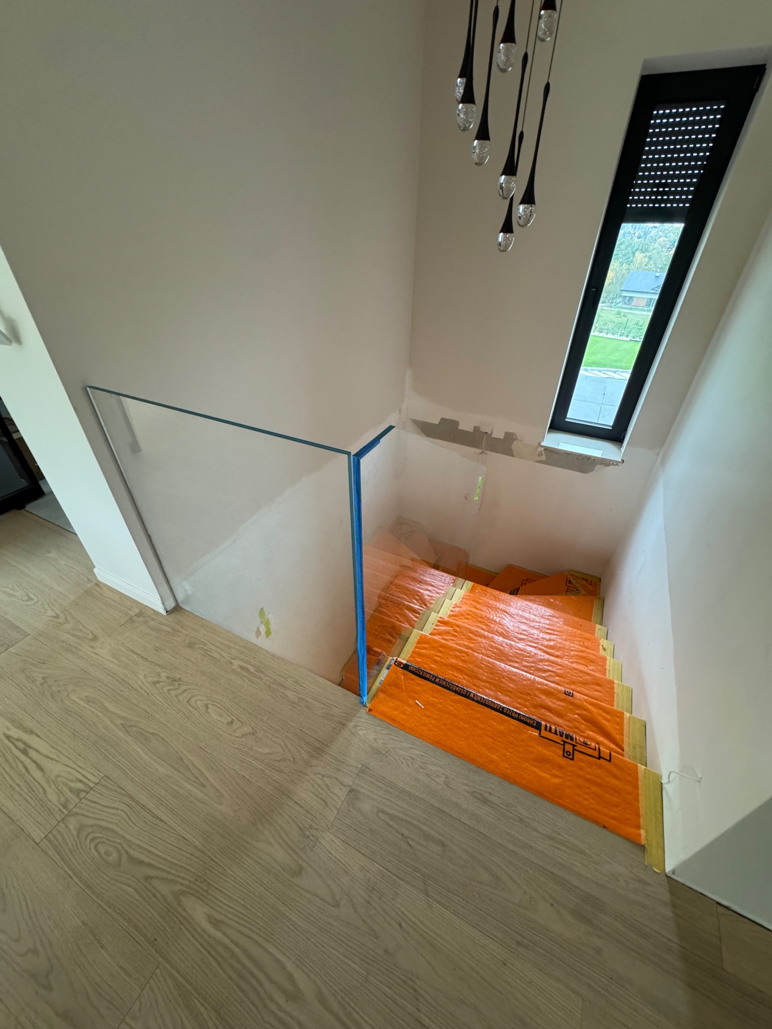 Szklana balustrada na schodach w trakcie montażu, zabezpieczone pomarańczową folią. Widoczne okno dachowe i lampa wisząca. Ujęcie z góry, jasne wnętrze.
