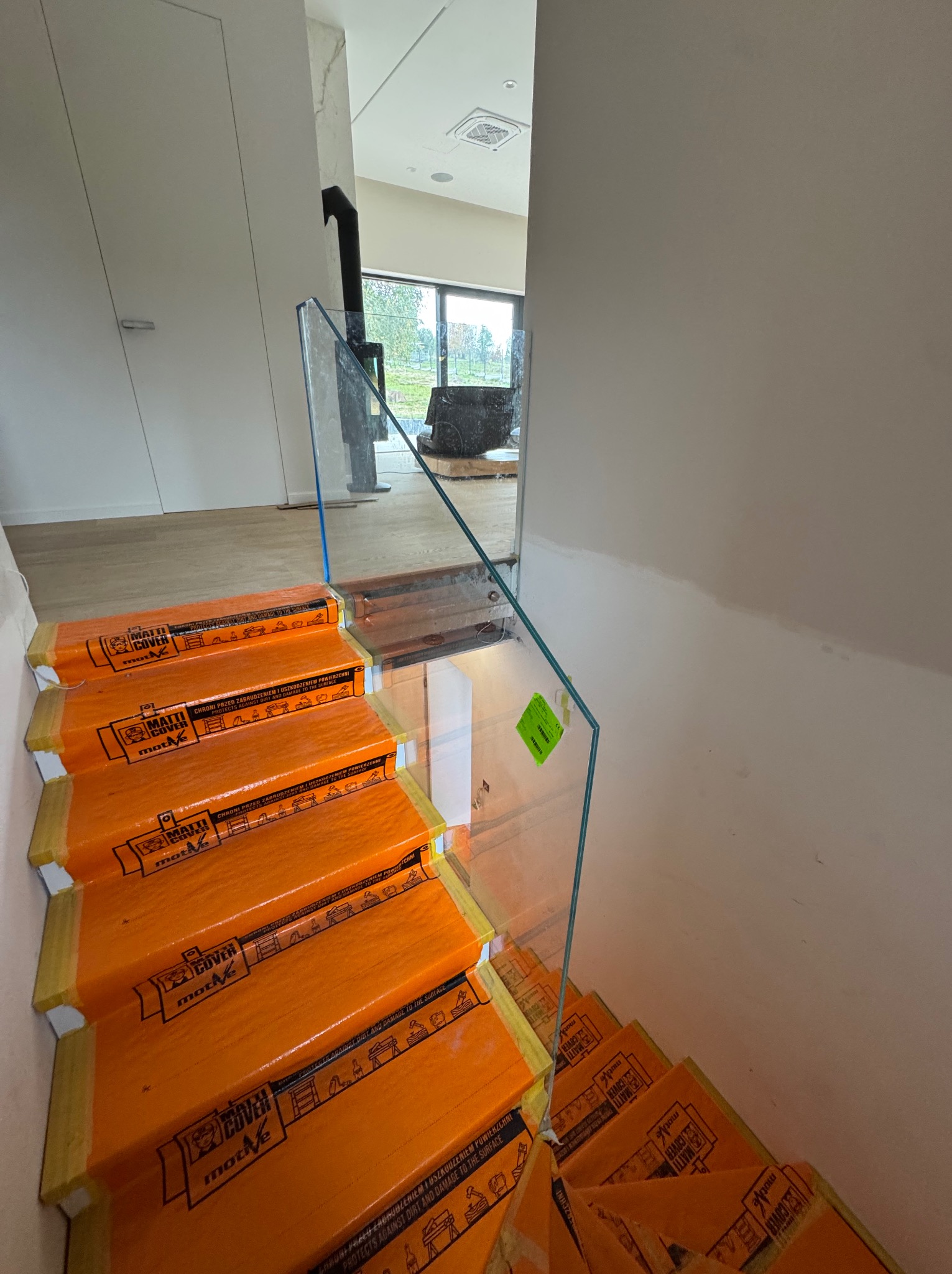 Szklana balustrada schodowa w trakcie montażu, zabezpieczona pomarańczową folią ochronną. Widok z góry na dół, wnętrze domu w tle.