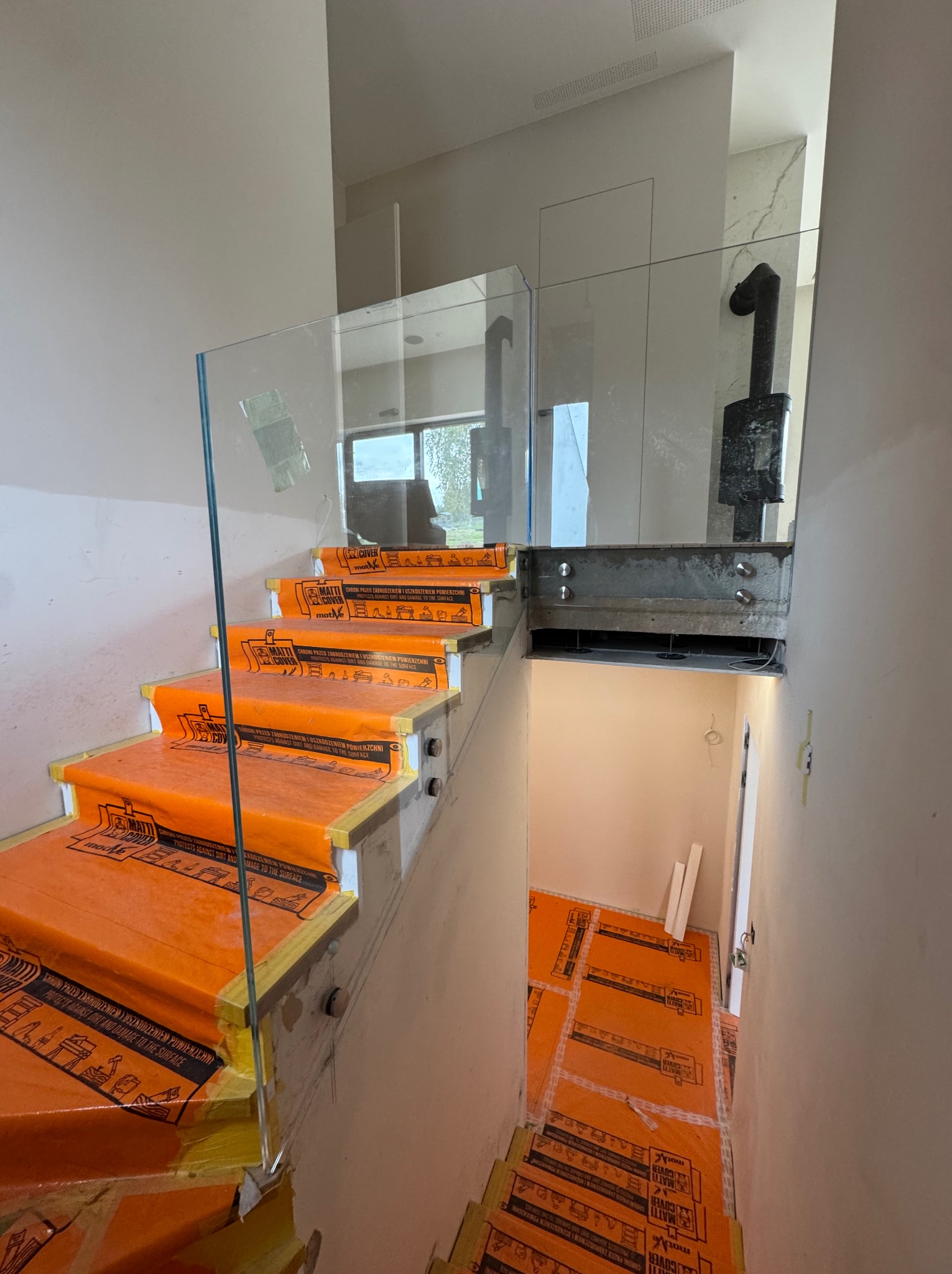 Szklana balustrada na schodach w trakcie montażu, zabezpieczone pomarańczową folią ochronną. Widoczne elementy mocujące i fragment wnętrza z kominkiem.