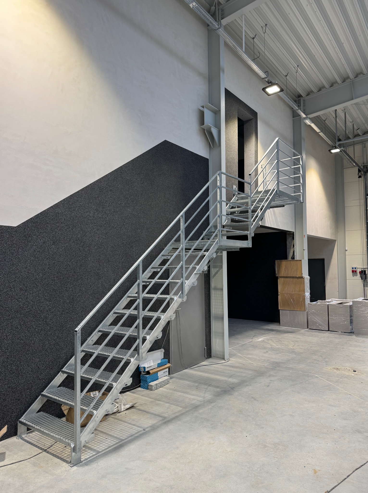 Metalowe schody kratowe z poręczami, prowadzące do otworu w ścianie w hali. Widoczne elementy konstrukcyjne budynku i materiały budowlane. Ujęcie z dołu, oświetlenie górne.