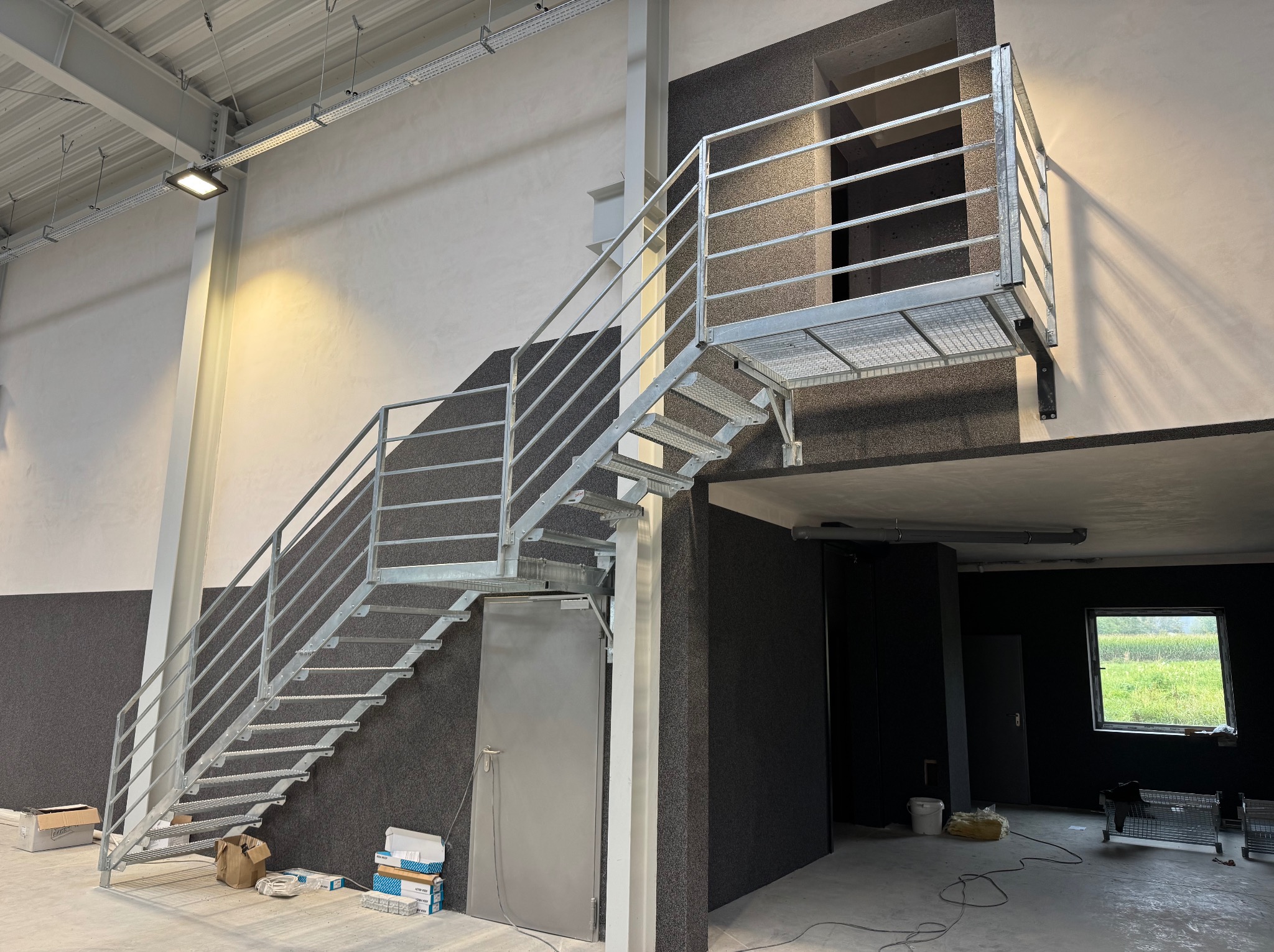 Metalowe schody z platformą na antresolę w hali, widok z dołu. Konstrukcja ocynkowana, z poręczami. W tle ściana z szarego tynku strukturalnego i drzwi.