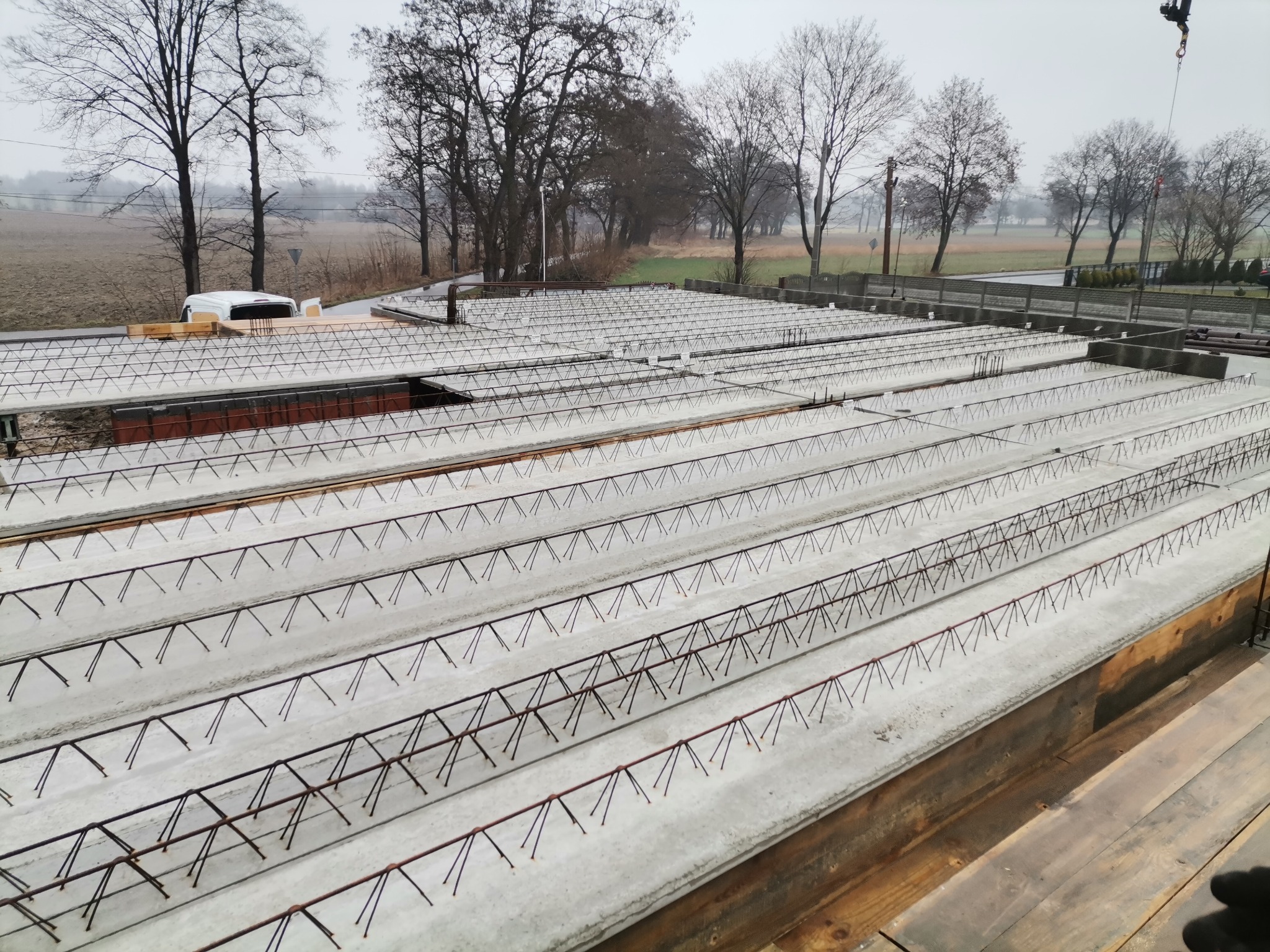 Zalany betonem strop Teriva z wystającymi prętami zbrojeniowymi, widok z góry na budowie domu jednorodzinnego, w tle drzewa i samochód dostawczy.