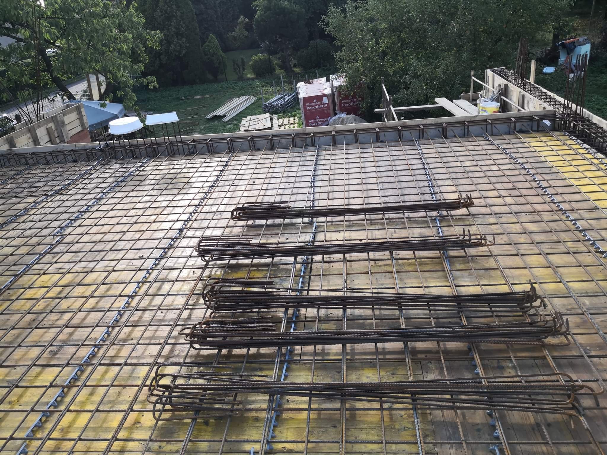 Przygotowanie zbrojenia stropu na deskowaniu z widocznymi prętami zbrojeniowymi, w tle materiały budowlane i zieleń.