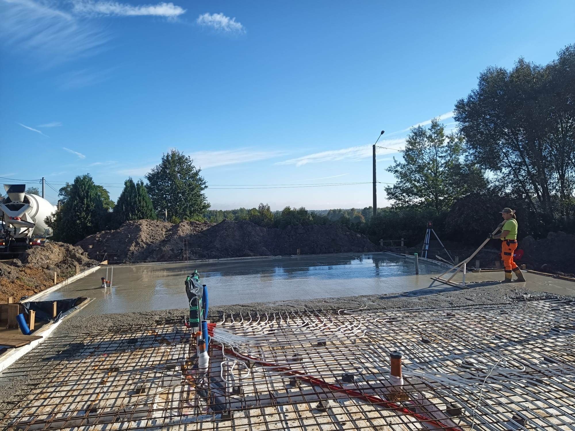 Wylewanie fundamentów pod dom jednorodzinny z widocznym zbrojeniem, instalacją ogrzewania podłogowego i pracownikiem wyrównującym beton łatą. W tle betonomieszarka i nasyp ziemi.