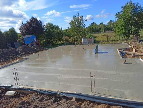 Wylany beton na przygotowanym fundamencie pod budowę domu, widoczne wystające pręty zbrojeniowe i rury instalacyjne, w tle domek dla dzieci na placu budowy.