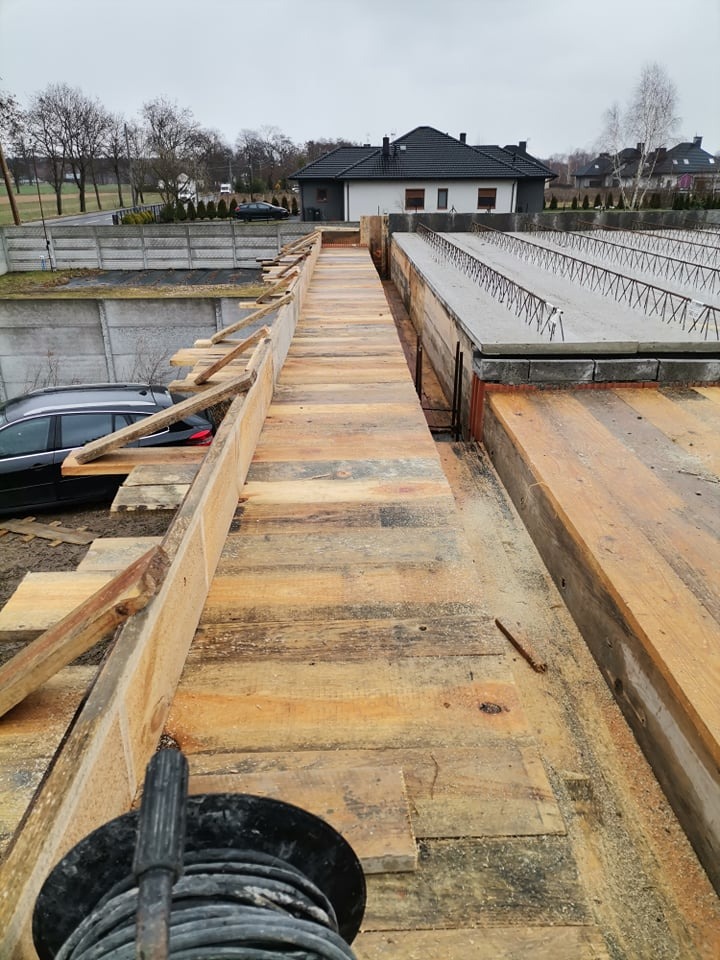 Szczegółowy widok szalunku i zbrojenia fundamentów domu jednorodzinnego w trakcie budowy, z widocznym kablem elektrycznym w dolnej części kadru i domem w tle.