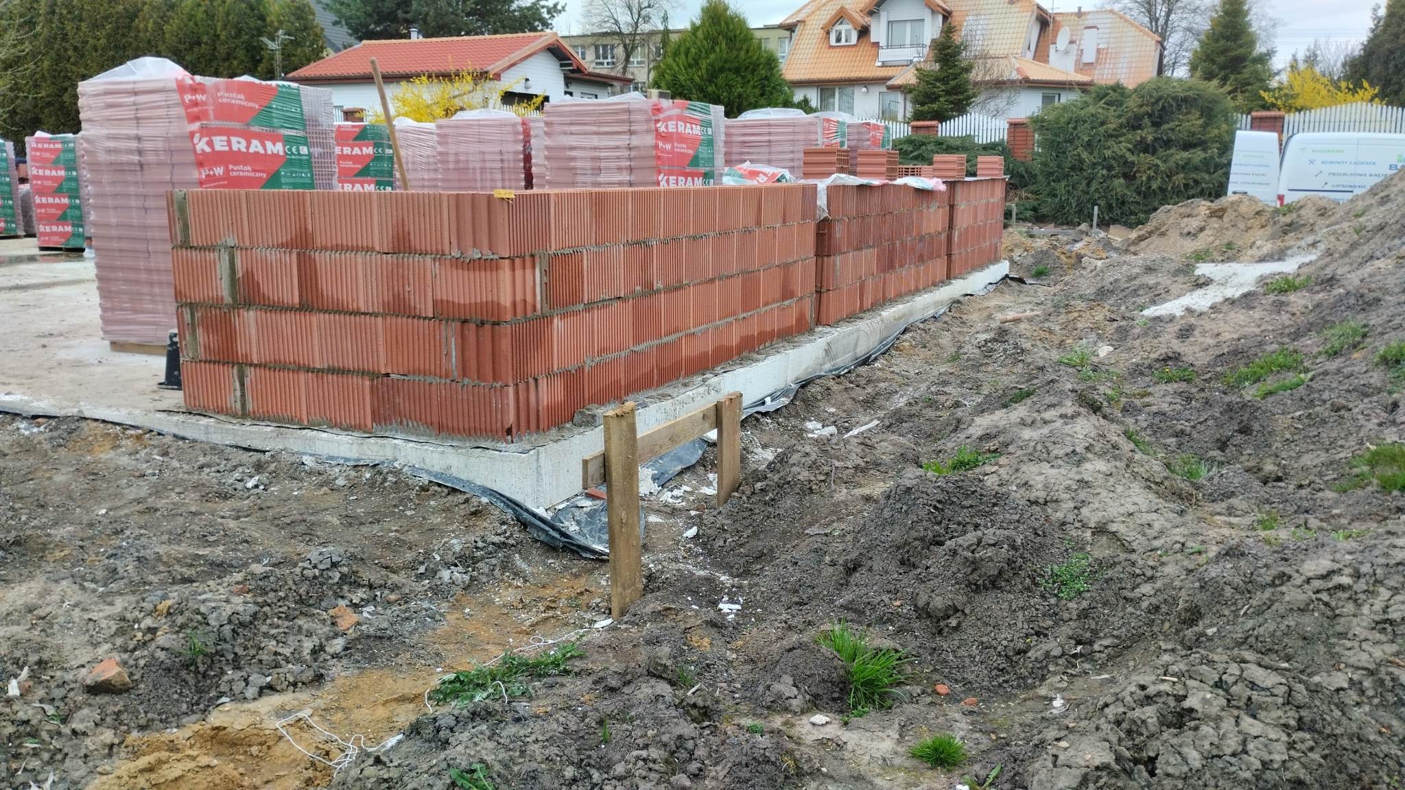Wznoszenie ścian parteru domu z czerwonych pustaków ceramicznych na betonowym fundamencie, widoczne zapasy pustaków w opakowaniach, teren budowy w trakcie prac ziemnych.