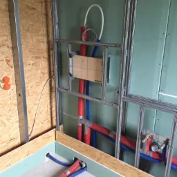 Wewnętrzna instalacja wodno-kanalizacyjna w trakcie montażu, widoczne rury czerwone i niebieskie, stelaż metalowy pod zabudowę oraz płyty kartonowo-gipsowe.