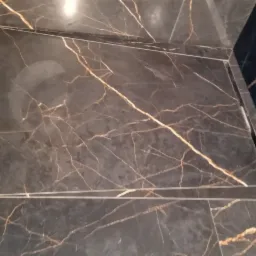 Ukończona kabina prysznicowa z czarnymi, marmurkowymi płytkami, widoczne detale wykończenia i listwy. Na ścianie po lewej stronie widoczne białe listwy z pomarańczową taśmą.