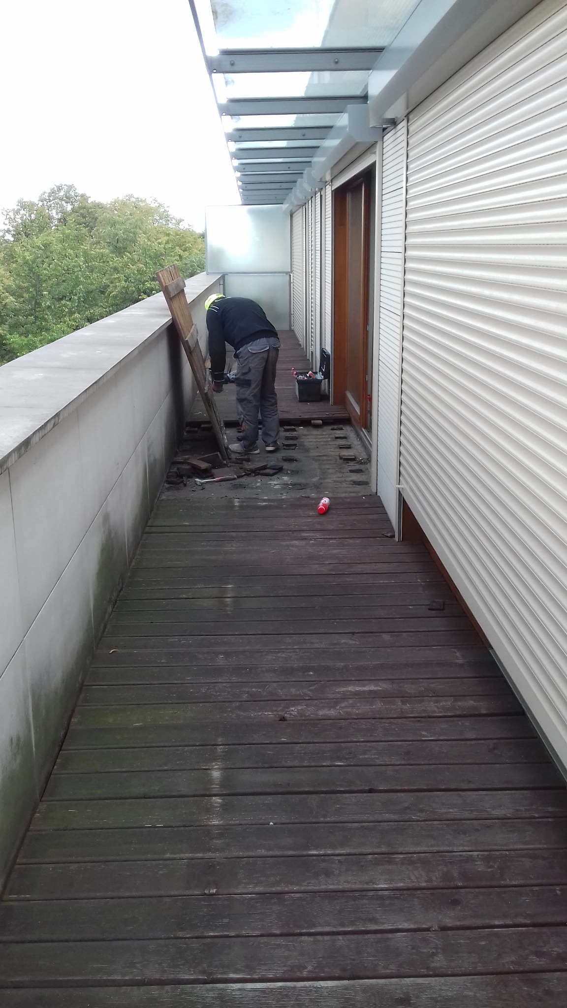 Remont drewnianego balkonu: pracownik w kasku usuwa stare deski, widoczna skrzynka z narzędziami i butelka na podłodze.