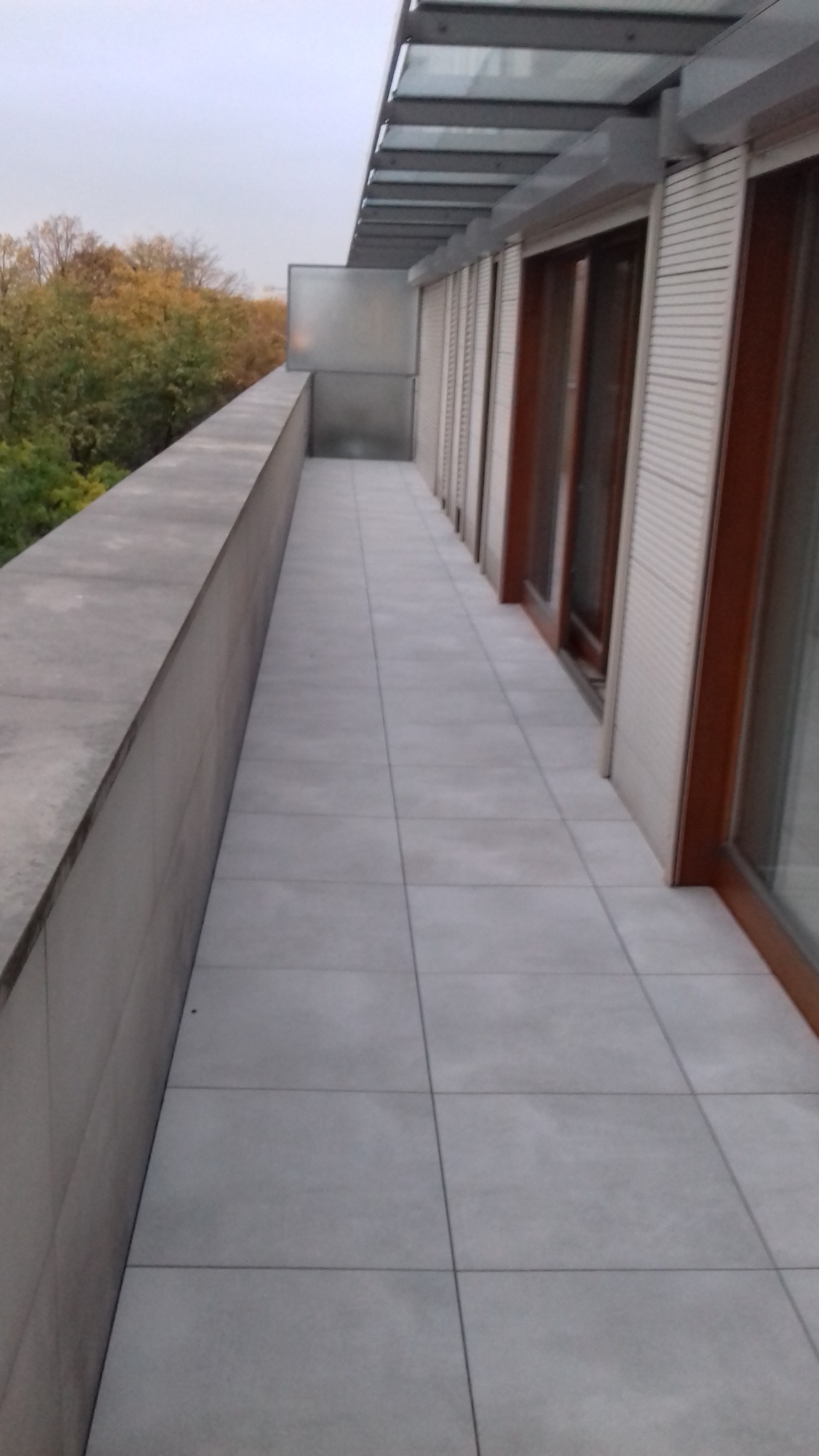 Długi balkon wyłożony szarymi płytkami, z widokiem na drzewa w jesiennych kolorach i szklanym zadaszeniem.