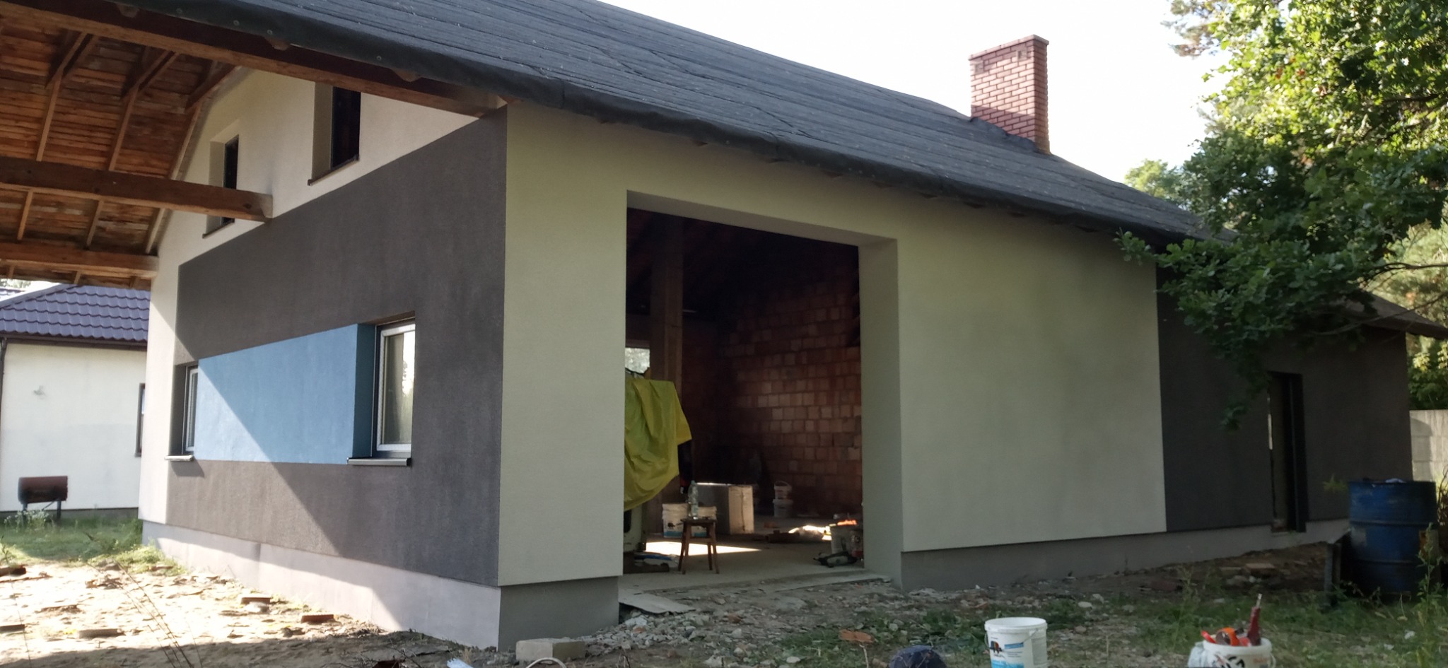 Dwukolorowa elewacja budynku z otwartym wjazdem do garażu, widoczne wiadro z farbą i narzędzia malarskie na zewnątrz, ceglana ściana wewnątrz garażu, żółta plandeka ochronna.