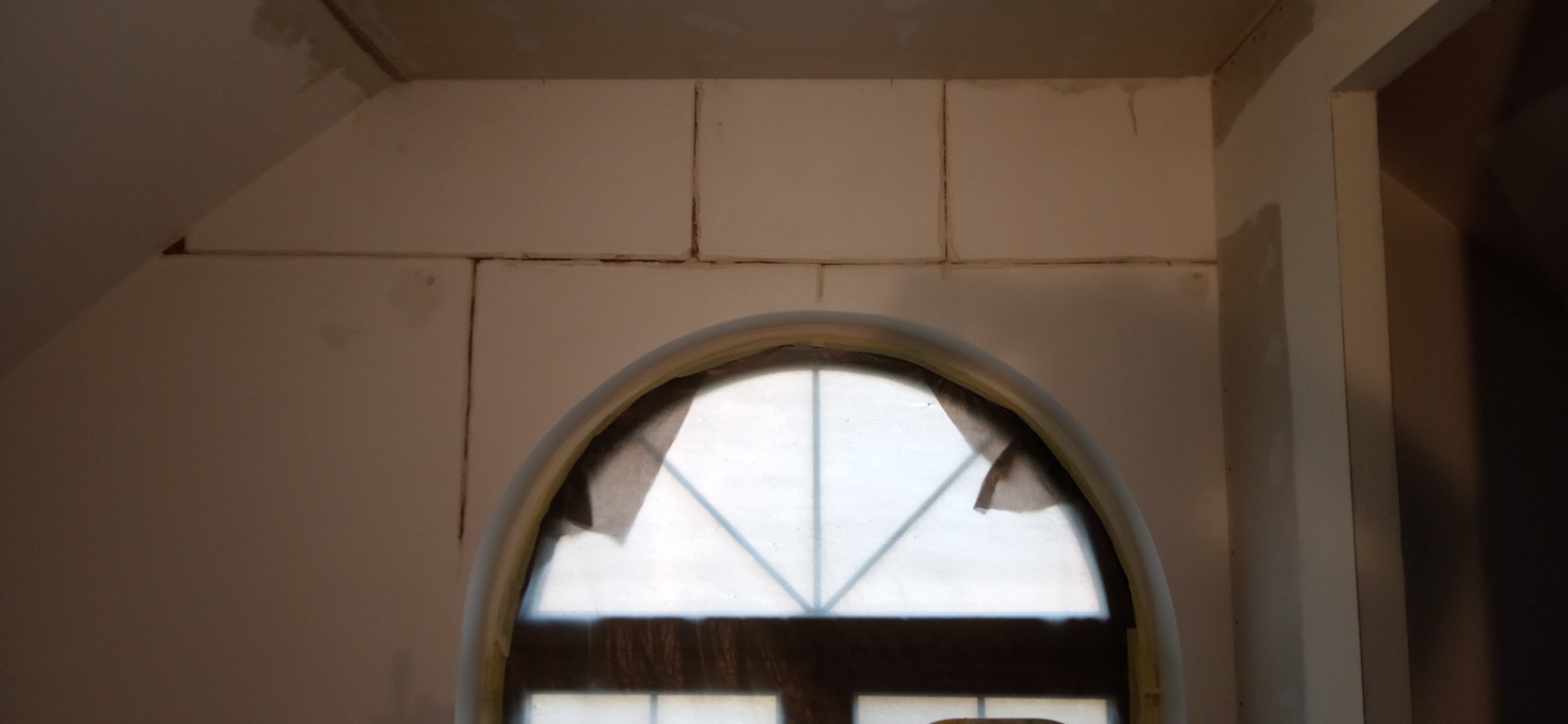 Montaż płyt kartonowo-gipsowych na poddaszu, widoczne łączenia płyt i okno łukowe zabezpieczone folią malarską.