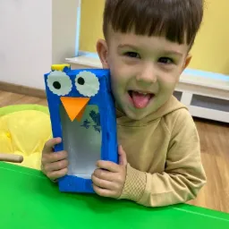 Uśmiechnięty chłopiec trzyma własnoręcznie wykonaną, niebieską zabawkę w kształcie pingwina z papieru, stojąc przy zielonym stole.