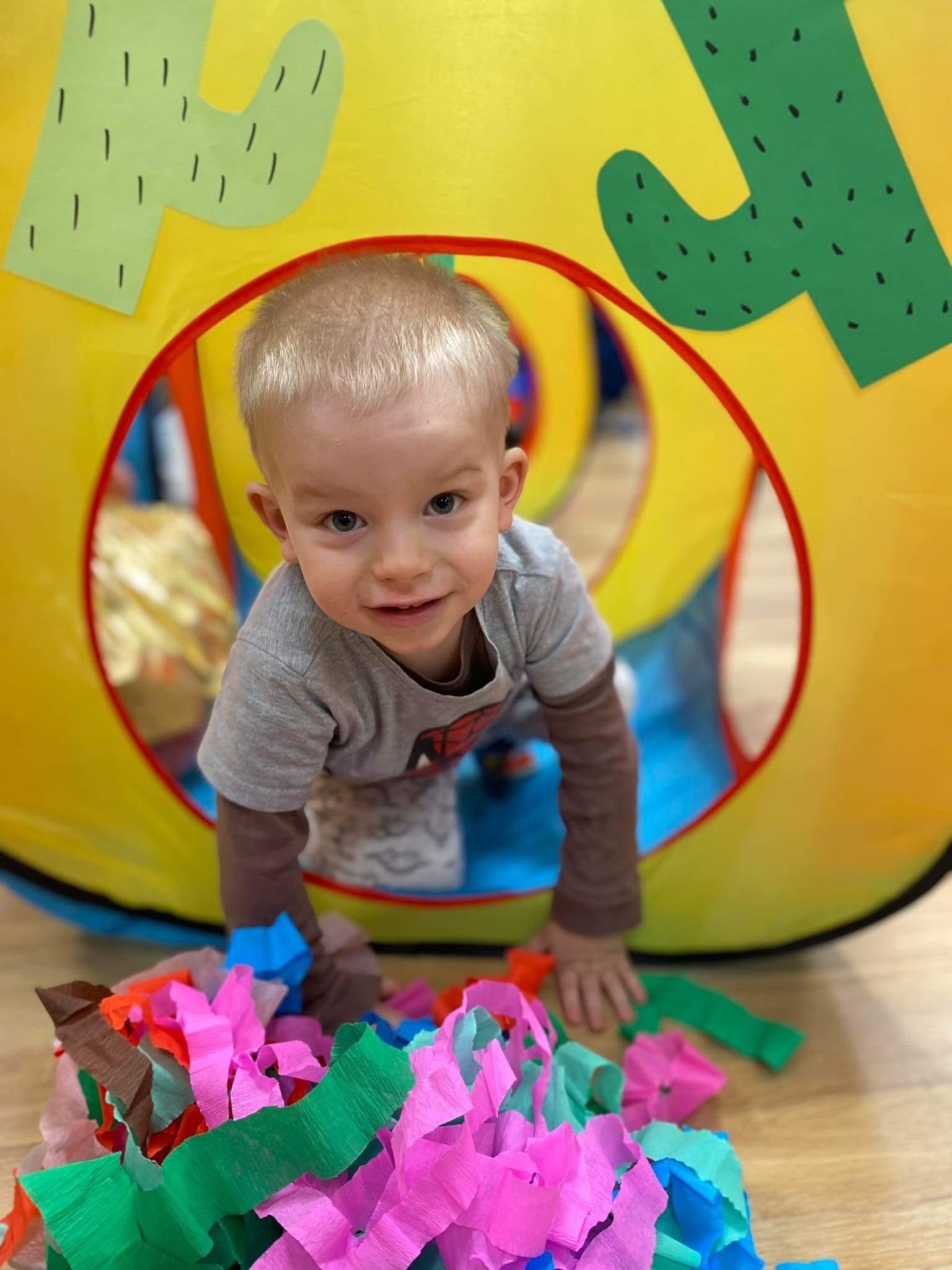 Uśmiechnięty blond chłopiec wychyla się z tunelu do zabawy z motywem kaktusa, w tle widoczna podłoga i kolorowa bibuła.