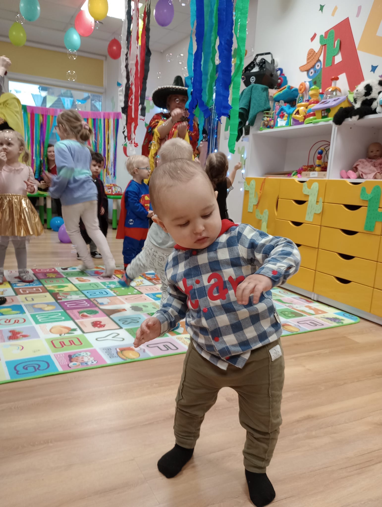 Dziecko w koszuli w kratę i spodniach khaki bawi się na kolorowej macie edukacyjnej w sali z dekoracjami imprezowymi, w tle animator w stroju meksykańskim puszcza bańki mydlane.