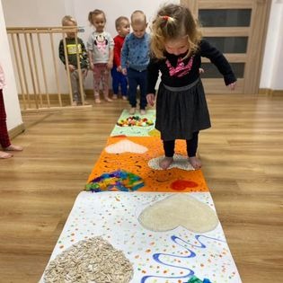 Dziewczynka w czarnej bluzce i szarej spódniczce przechodzi boso po sensorycznej ścieżce z różnymi fakturami, za nią stoi grupka dzieci w różnym wieku.