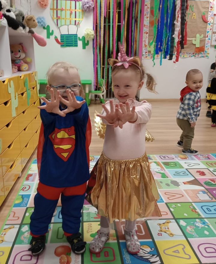 Dwoje uśmiechniętych dzieci w kostiumach - chłopiec w stroju Supermana i dziewczynka z jednorożcem na głowie i złotą spódniczką - stoją na kolorowej macie w sali z dekoracjami.