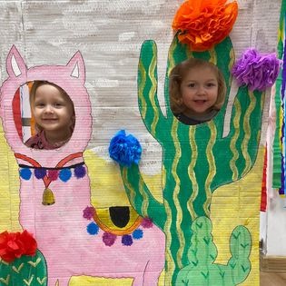 Dwie uśmiechnięte dziewczynki pozują za kartonową dekoracją: jedna w otworze w kształcie lamy w różowym kostiumie, druga w otworze w kształcie kaktusa ozdobionego kolorowymi pomponami.