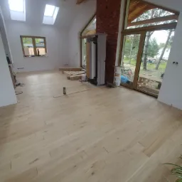 Jasna, drewniana podłoga ułożona w jodełkę francuską w przestronnym pokoju z dużymi oknami i ceglanym kominem. Widoczne deski podłogowe przygotowane do montażu oraz poziomnica.