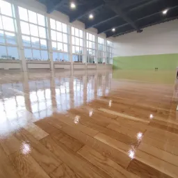 Błyszcząca, nowo polakierowana drewniana podłoga w hali sportowej w Warszawie, z widocznymi refleksami światła odbijającymi się od gładkiej powierzchni.