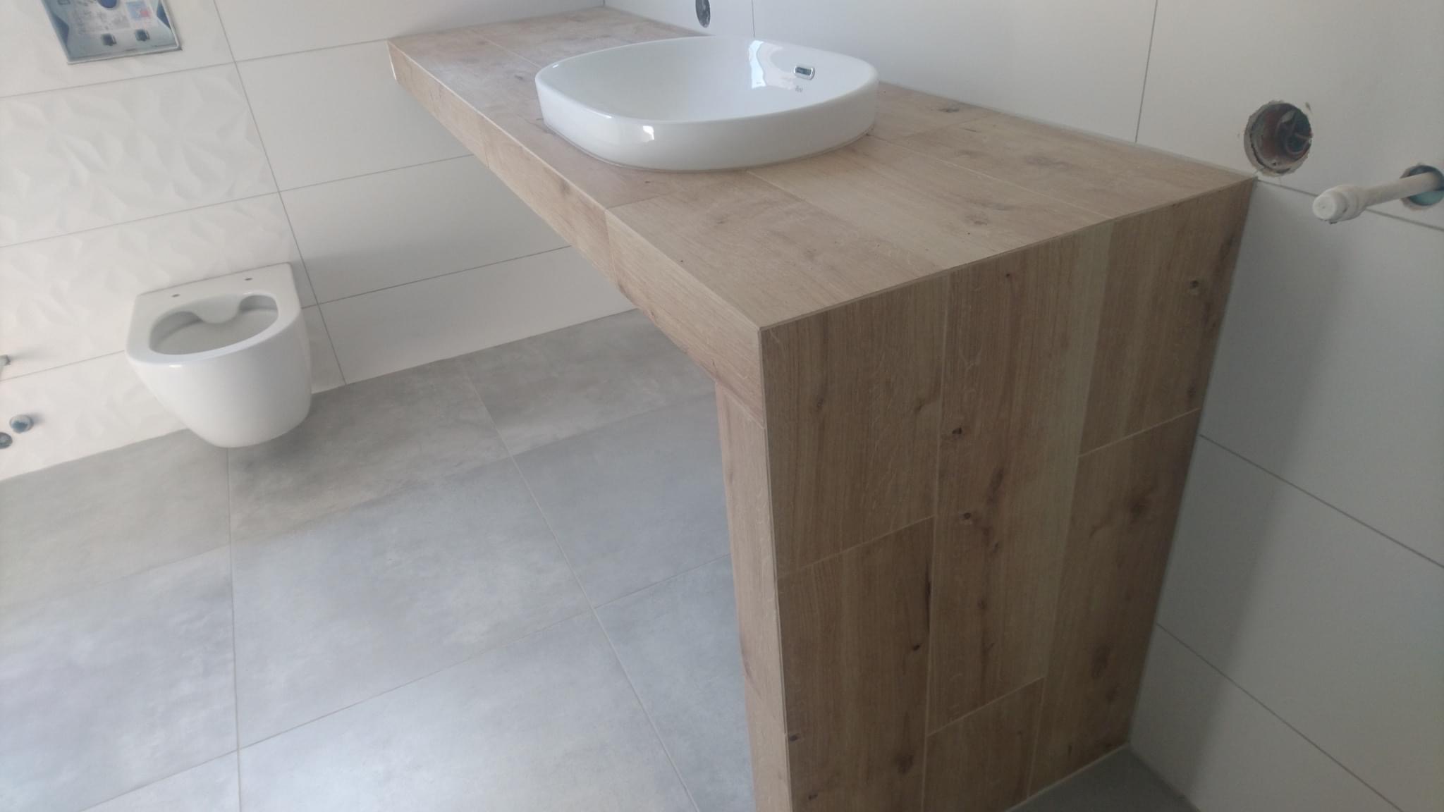 Nowoczesna łazienka z podwieszaną toaletą i umywalką nablatową na blacie imitującym drewno, szare płytki podłogowe i białe ścienne z delikatnym wzorem.