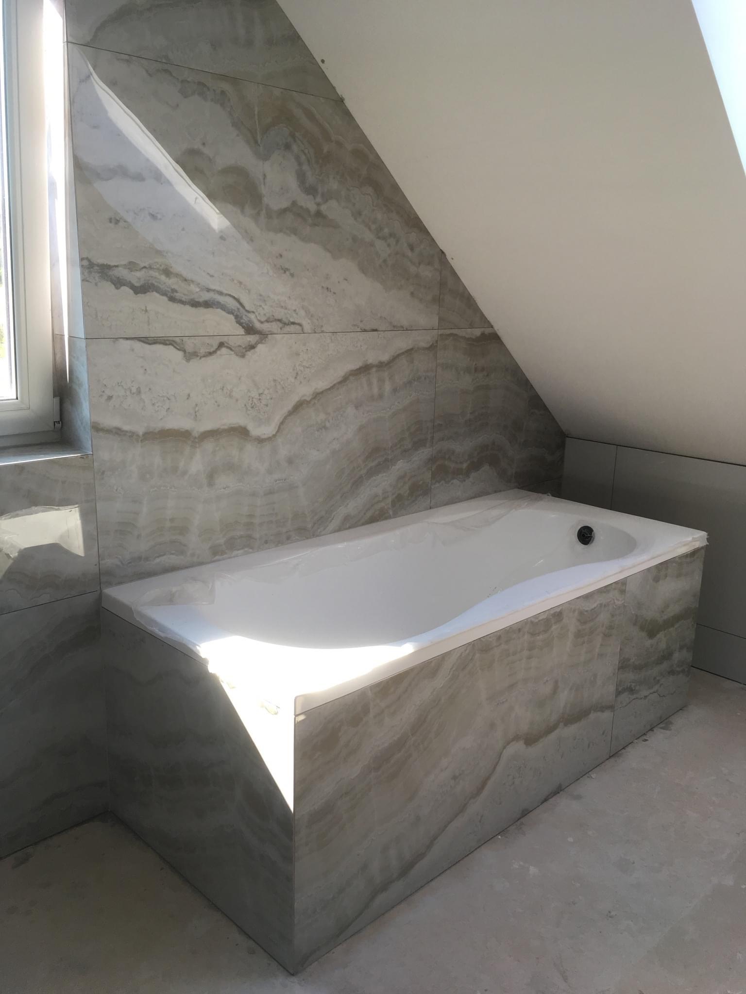 Wanna obudowana płytkami imitującymi onyks w łazience na poddaszu, widoczne okno i surowy beton na podłodze.