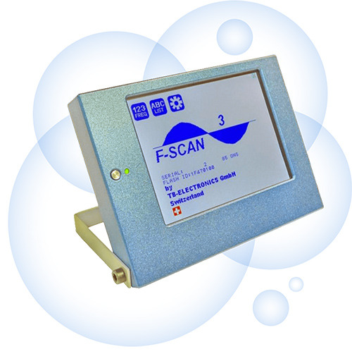 Urządzenie F-SCAN z ekranem wyświetlającym sinusoidę, obudowa w kolorze jasnoniebieskim, widoczna dioda LED i dane producenta TB-ELECTRONICS GmbH ze Szwajcarii.