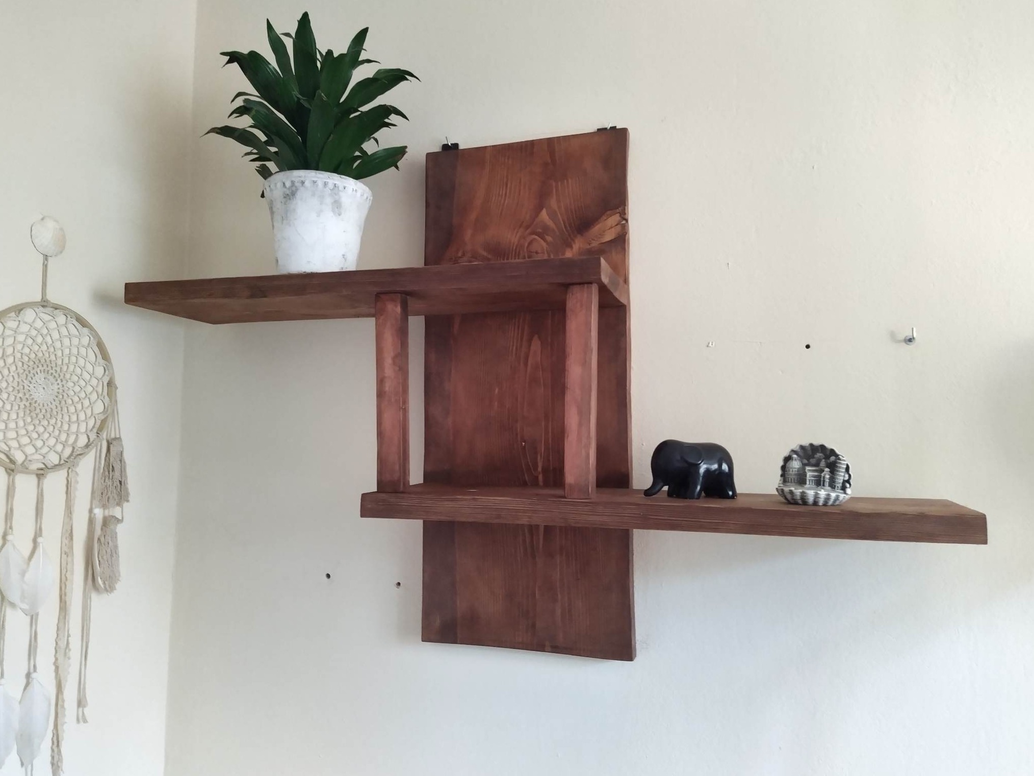 Drewniana półka ścienna o asymetrycznym kształcie, na której stoją doniczka z zieloną rośliną, figurka słonia i pamiątka z Włoch, obok wisi łapacz snów